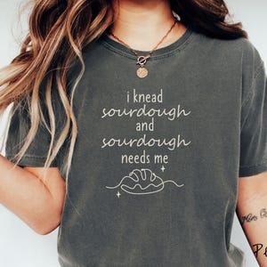May include: A dark green t-shirt with the text "i knead sourdough and sourdough needs me" in white. A simple line drawing of a loaf of bread is below the text.