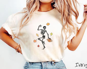 May include: Ivory-colored t-shirt featuring a black skeleton playing football, surrounded by falling autumn leaves in orange, yellow, and green. The skeleton is holding a brown football. The word "Ivory" is written in black script.