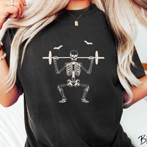 May include: Black t-shirt featuring a white skeleton lifting weights, with two bats above. The word "Black" is written in a script font at the bottom right. A fun, spooky design for Halloween or fitness enthusiasts.