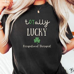 May include: Black t-shirt with the words "totally LUCKY" in white and green, a green shamrock, and the text "Occupational Therapist." The shirt is a crew neck style.
