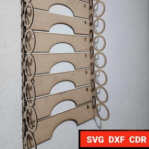 Puede incluir: Un organizador de pared de madera con ocho ranuras para guardar objetos. El organizador tiene un acabado marrón claro y está hecho de madera contrachapada. Las ranuras tienen forma de arco y están diseñadas para guardar objetos como gafas, sombreros o bufandas.