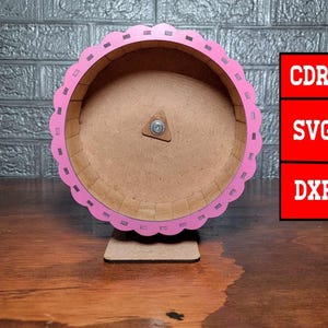 Puede incluir: Rueda de hámster de madera rosa con un soporte. La rueda tiene un borde festoneado y un interior marrón. El soporte es de madera y del mismo color que la rueda.
