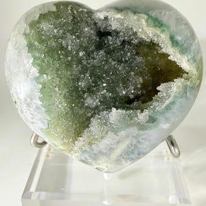 May include: A heart-shaped geode with a hollow center filled with sparkling crystals. The geode has a green and white exterior and is displayed on a clear acrylic stand. A unique decorative item.