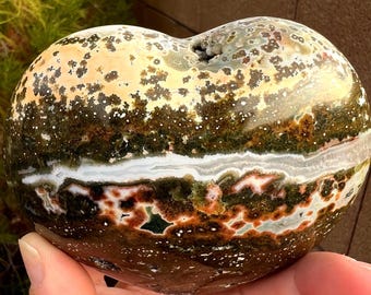 May include: A heart-shaped stone with a unique pattern of colors. The stone features a mix of beige, brown, green, and white hues, with a speckled texture. The stone is held in a hand, showcasing its natural beauty and intricate design.