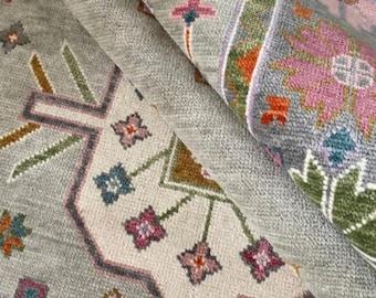 May include: A rolled-up, intricately patterned rug with a muted color palette. The design features floral and geometric motifs in shades of gray, pink, green, and orange. The rug's texture appears soft and plush, suggesting a comfortable floor covering.