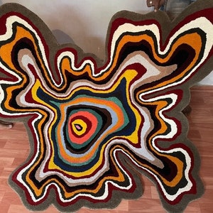 Op de afbeelding: Een kleurrijk abstract tapijt met een draaiend, gelaagd ontwerp in tinten rood, oranje, geel, groen, blauw en bruin. Het tapijt heeft een unieke, organische vorm.
