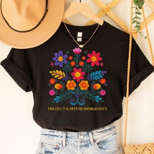 Puede incluir: Camiseta negra con un dise&ntilde;o floral colorido y el texto "Protect & Defend Immigrants". La camiseta est&aacute; en una percha con un sombrero de paja y una bolsa tejida.