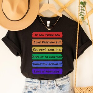 May include: Black t-shirt with a rainbow stripe design and the text "If you think you love freedom but you don't care if it applies to everyone what you actually love is privilege."