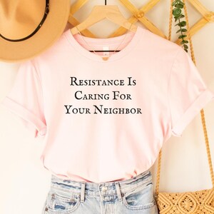 May include: A pink t-shirt with the text "Resistance is Caring For Your Neighbor" on a white background. The shirt is hanging on a wooden hanger with a straw hat and a woven bag.