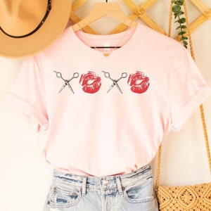 May include: A pink t-shirt with a graphic of scissors and red lips. The shirt is hanging on a wooden hanger and is styled with denim shorts and a woven bag.
