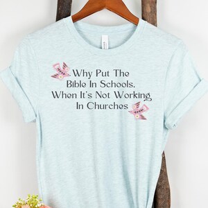 May include: A light blue t-shirt with the text "Why Put The Bible In Schools, When It's Not Working In Churches" printed on the front. The text is in black and there are two pink birds on either side of the text.