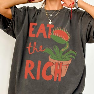 May include: A charcoal gray short-sleeve t-shirt featuring the red text EAT the RICH and a graphic of a green Venus flytrap plant in a terracotta-colored pot. The shirt has a crew neckline and a relaxed fit, showcasing a screen-printed design.