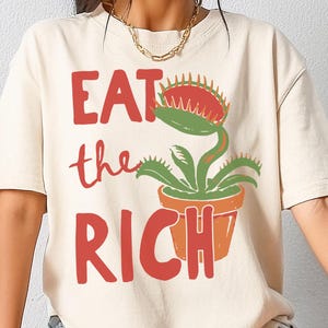 May include: A cream-colored short-sleeve t-shirt featuring a graphic of a green Venus flytrap in an orange pot. The text "EAT the RICH" is printed in large red letters to the left of the plant illustration on the front of the garment.