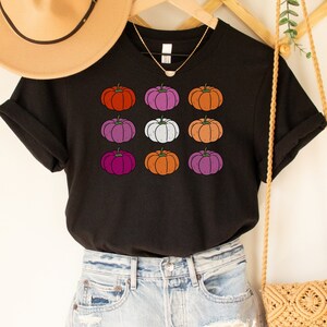 May include: A black t-shirt with a graphic of a grid of colorful pumpkins. The shirt is styled with a straw hat, denim shorts, and a crocheted bag.