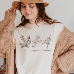May include: A white t-shirt with a graphic of three different types of trees: fir, dogwood, and tamarack. The trees are illustrated in a vintage style. The shirt is being worn by a person with brown hair and a brown hat.