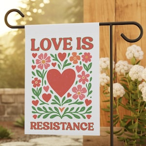 May include: A white garden flag with the words "LOVE IS RESISTANCE" in red, with a large red heart and floral design. The flag is displayed on a black metal stand in a garden setting.