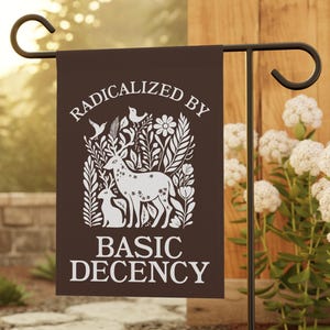 May include: A brown garden flag with the text "RADICALIZED BY BASIC DECENCY" in white. The flag features a white illustration of a deer, a fawn, birds, and foliage. The flag is displayed on a black metal stand in a garden setting.