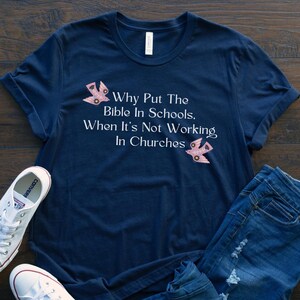 May include: A navy blue t-shirt with white text that reads "Why Put The Bible In Schools. When It's Not Working In Churches". The shirt has two pink bird graphics on it. The shirt is laid out on a wooden surface with a pair of white Converse shoes and blue jeans.