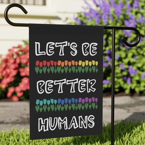 May include: A black garden flag with the text "LET'S BE BETTER HUMANS" in white. The flag features rows of colorful tulips in red, orange, yellow, green, blue, and purple. The flag is displayed on a black metal stand in a grassy yard.