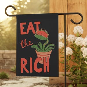 May include: A black rectangular garden flag featuring the red text EAT the RICH and a green Venus flytrap plant in a terracotta-colored pot. The flag is displayed on a black metal stand, positioned vertically with the text clearly visible.
