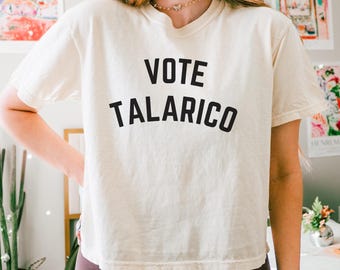 May include: Cream-colored t-shirt with the words "VOTE TALARICO" printed in bold black letters. The shirt is a short-sleeved, casual style. The person is wearing dark brown leggings.