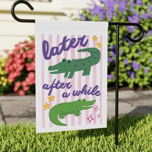 May include: A rectangular garden flag featuring a white background with vertical light pink stripes. The text "later after a while" is written in purple script. Two green crocodiles are illustrated, accompanied by yellow stars and pink sparkles.