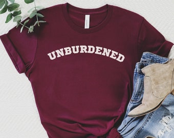 May include: A maroon t-shirt with the word "UNBURDENED" printed in white on the front. The shirt is styled with blue jeans and brown ankle boots.
