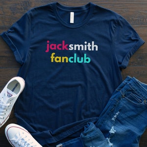 May include: A navy blue t-shirt with the text "jacksmith fanclub" in colorful letters. The shirt is laid flat on a wooden surface with a pair of white sneakers and blue jeans.