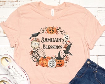 May include: A peach-colored t-shirt with a graphic design of a wreath with pumpkins, crows, and a clock. The text on the shirt reads "Samhain Blessings".