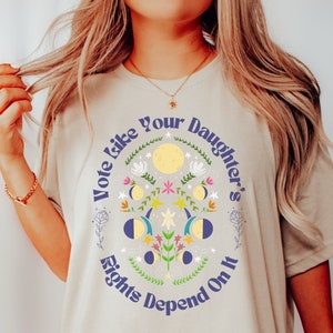 May include: A light beige t-shirt with a graphic print of a circle with the text "Vote Like Your Daughter's Rights Depend On It" and a floral design with moons and stars.