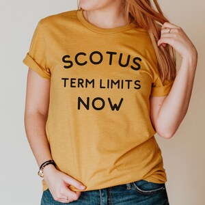 May include: A mustard yellow t-shirt with black text that reads "SCOTUS TERM LIMITS NOW".