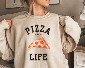 Könnte beinhalten: Beiges Sweatshirt mit dem schwarzen Schriftzug "PIZZA is LIFE". Unter dem Wort "is" befindet sich eine Aquarell-Illustration eines Pizzastücks mit Peperoni. Das Sweatshirt hat eine neutrale Farbe und scheint aus einem weichen Material zu sein.