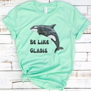 May include: A light green t-shirt with a black and white orca graphic and the text "Be Like Gladys".