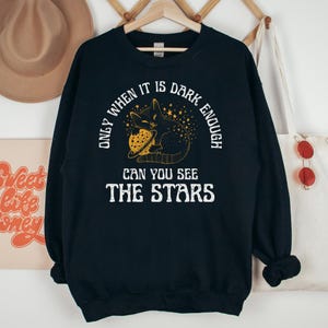 May include: Black sweatshirt with a black cat holding a planet and stars. The text reads "Only when it is dark enough can you see the stars."