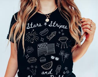 Op de afbeelding: Zwart T-shirt met witte illustraties van vuurwerk, een hotdog en een Amerikaanse vlag. De tekst "Stars Stripes and Human Rights" is in witte letters geschreven, samen met andere patriottische symbolen.
