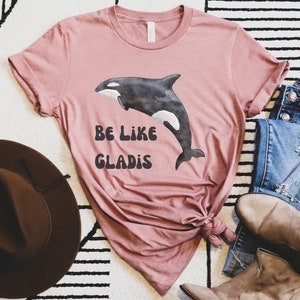 May include: A pink t-shirt with a black and white watercolor illustration of an orca whale. The shirt has the text "Be Like Gladys" printed on it in black.