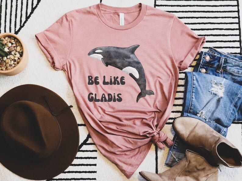 May include: A pink t-shirt with a black and white watercolor illustration of an orca whale. The shirt has the text "Be Like Gladys" printed on it in black.