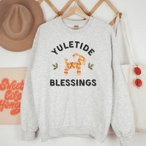 May include: A light gray sweatshirt with the words "YULETIDE BLESSINGS" in black. A decorative Dala horse graphic is in the center, with holly accents. A tan hat and a tote bag are visible in the background.