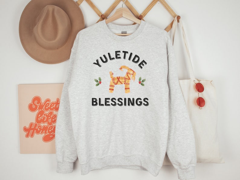 May include: A light gray sweatshirt with the words "YULETIDE BLESSINGS" in black. A decorative Dala horse graphic is in the center, with holly accents. A tan hat and a tote bag are visible in the background.