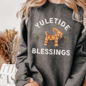 May include: Dark gray sweatshirt with the text "YULETIDE BLESSINGS" in white. The design includes a straw goat and holly sprigs. The sweatshirt is a comfortable, casual style, perfect for the holiday season.
