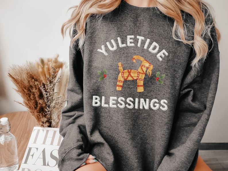 May include: Dark gray sweatshirt with the text "YULETIDE BLESSINGS" in white. The design includes a straw goat and holly sprigs. The sweatshirt is a comfortable, casual style, perfect for the holiday season.