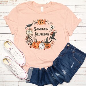 May include: A peach-colored t-shirt with a graphic design of a wreath with pumpkins, crows, and a clock. The text on the shirt reads "Samhain Blessings".