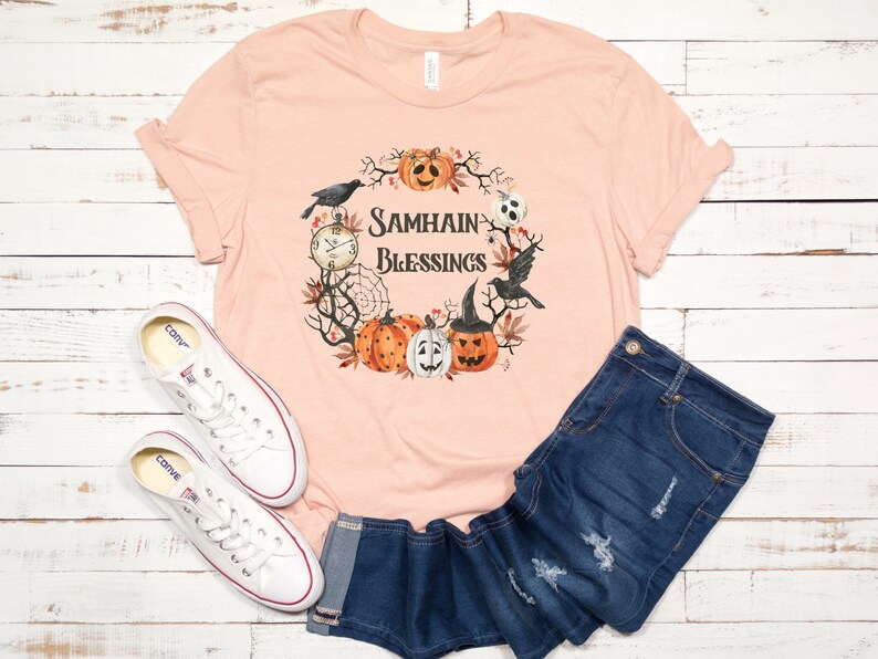 May include: A peach-colored t-shirt with a graphic design of a wreath with pumpkins, crows, and a clock. The text on the shirt reads "Samhain Blessings".