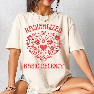 May include: Cream-colored t-shirt with the text "RADICALIZED BY BASIC DECENCY" in red. The design features a heart-shaped floral arrangement in red. The shirt has short sleeves and a relaxed fit.