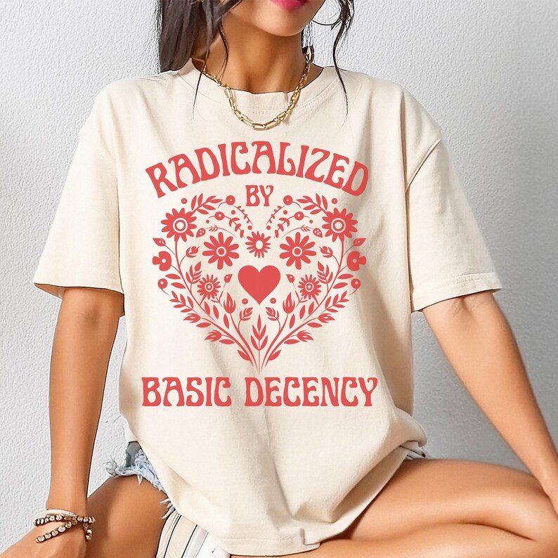May include: Cream-colored t-shirt with the text "RADICALIZED BY BASIC DECENCY" in red. The design features a heart-shaped floral arrangement in red. The shirt has short sleeves and a relaxed fit.