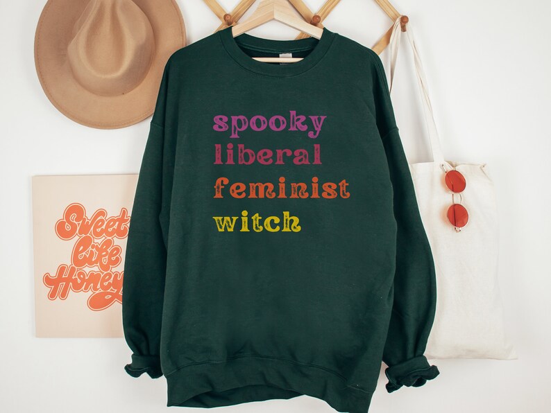 May include: Dark green sweatshirt with the text "spooky liberal feminist witch" in rainbow colors.