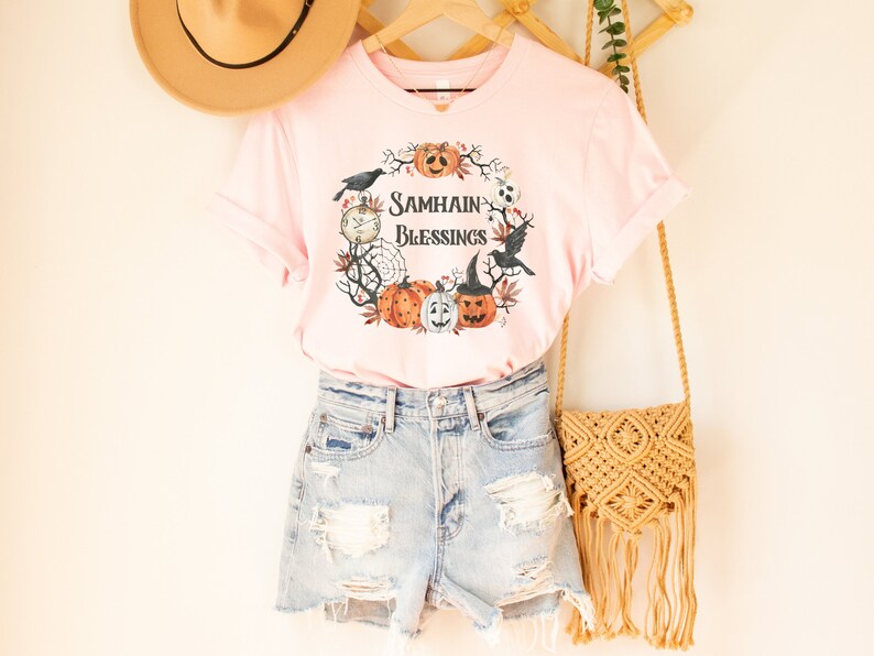 May include: Pink t-shirt with a graphic of a wreath with pumpkins, crows, and the text "Samhain Blessings".