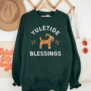 May include: Dark green sweatshirt with the words "YULETIDE BLESSINGS" in white. The design includes a straw goat and holly sprigs. The sweatshirt is hanging on a wooden hanger.