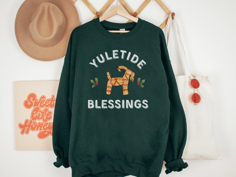 May include: Dark green sweatshirt with the words "YULETIDE BLESSINGS" in white. The design includes a straw goat and holly sprigs. The sweatshirt is hanging on a wooden hanger.