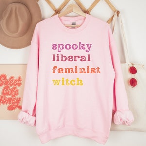 May include: Pink sweatshirt with the text "spooky liberal feminist witch" in rainbow colors.